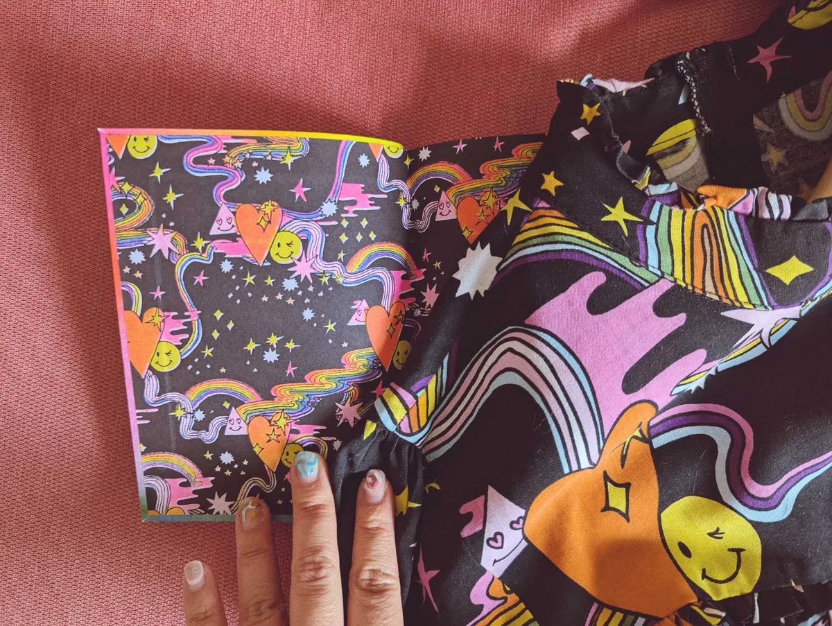 The opening spread of a book with a colorful, psychedelic pattern that matches the dress that is laying on top of the book.