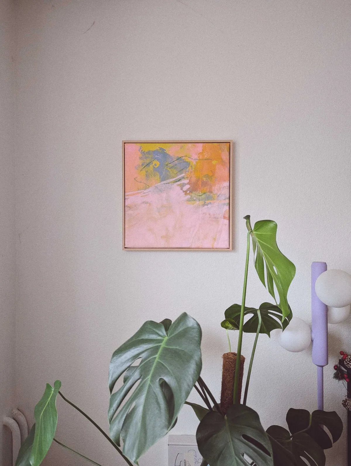 A square, colorful, abstract painting hangs on the wall, above a monstera plant and next to a modern, lavender floor lamp.