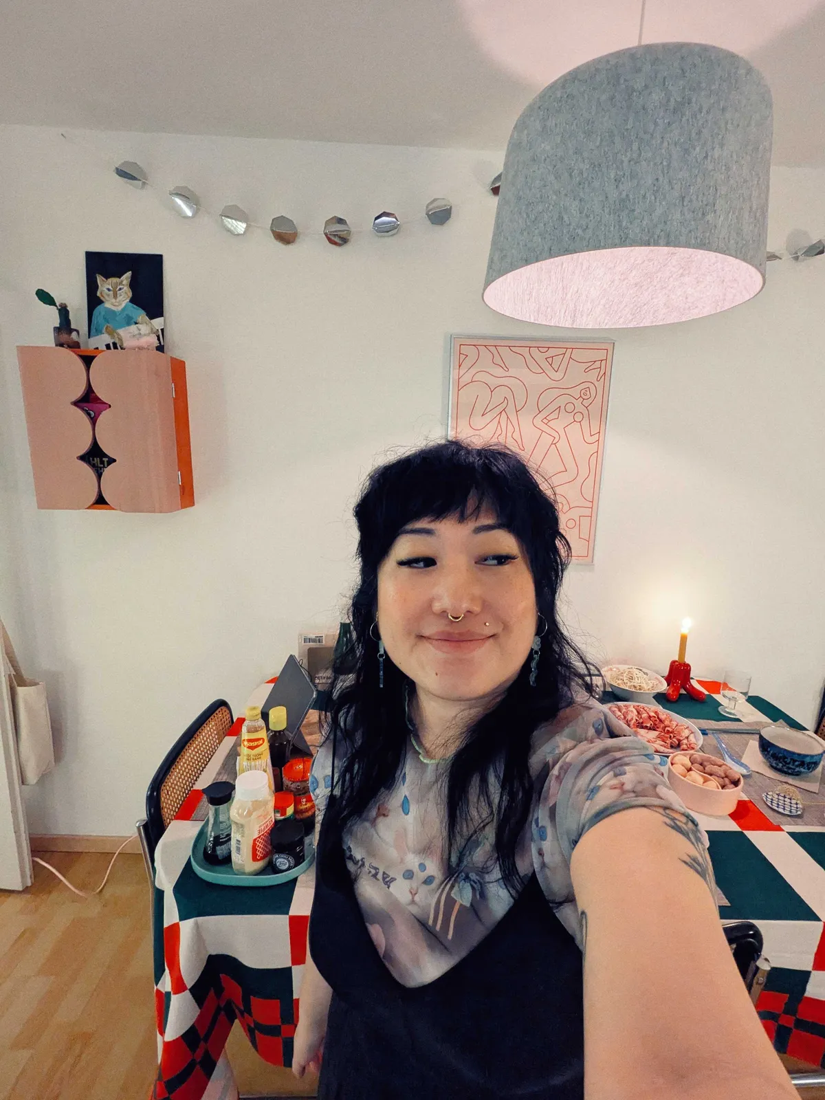 Peige taking a selfie, looking off to the side while smiling. The table is filled with food, and a chain of silver-colored circles hangs from the wall.