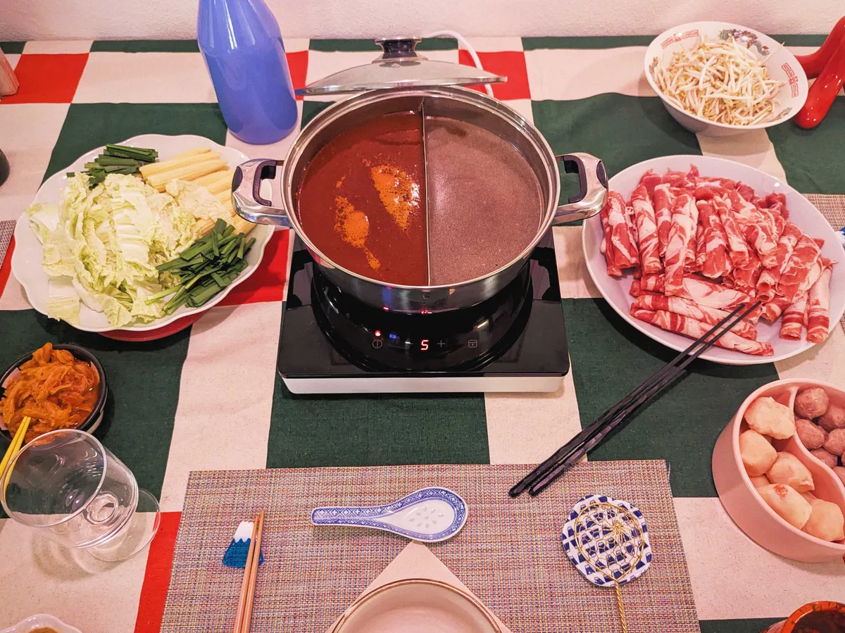 A table spread consisting of a hot pot with two types of soups (centered), a plate of veggies on the left, and plates of meat and meatballs on the right.