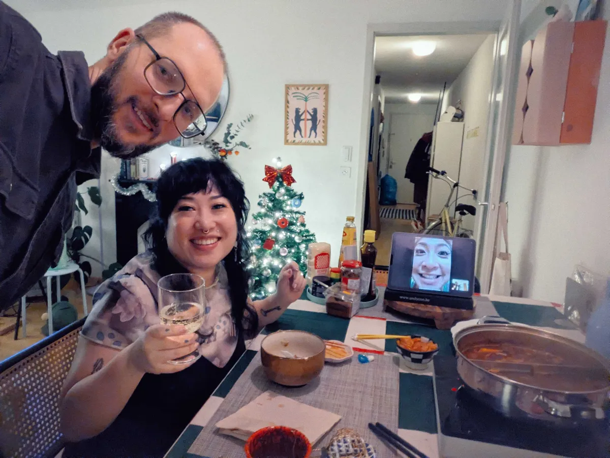 Peige is sitting on a chair, smiling and "toasting" the camera with a glass of wine. Daniel is standing, his body cropped out, also smiling. Peige's sister is smiling, her entire face filling the screen, from the iPad at the end of the table.