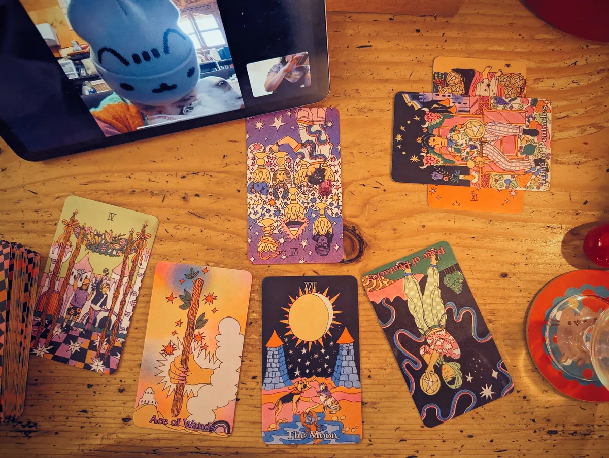 An overhead view of colorful tarot cards, seemingly organized like a spread. Peige's sister seen on the iPad, wearing a Pusheen beanie and sniffing her dog.