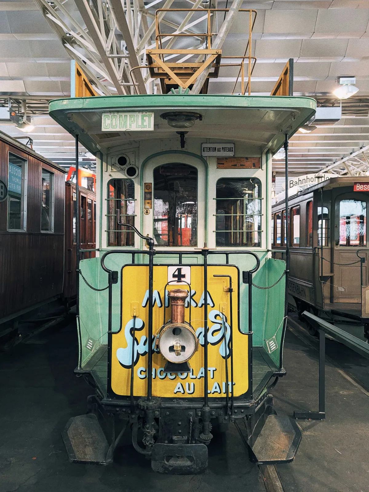 A cute seafoam green colored tram with a yellow door sits idly amongst other trams. The words "chocolat au lait" is hand-painted on the door, with some other words in script.