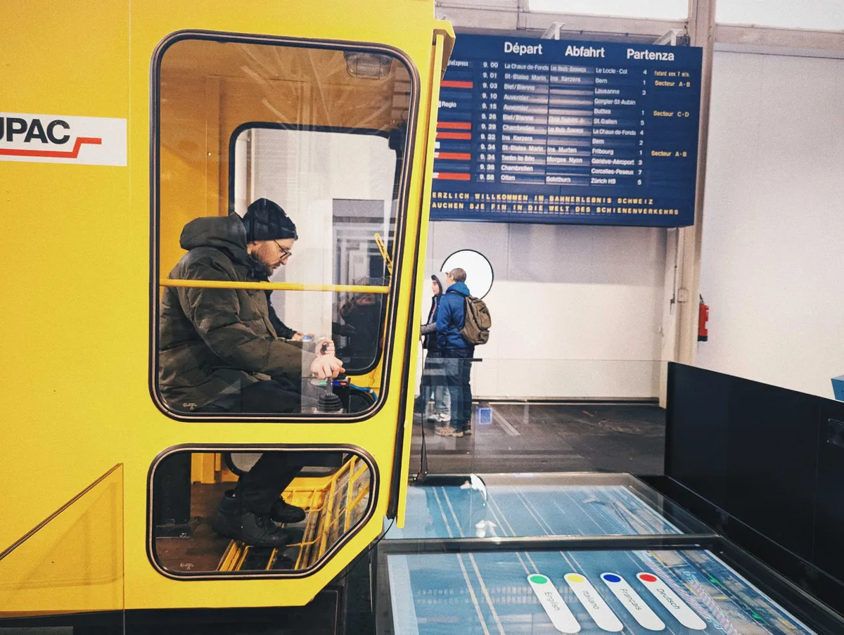 Daniel sits inside a yellow vehicle with his hands on some knobs and dials. He is staring at a giant screen below.