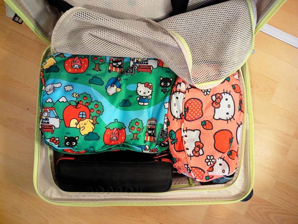 An open, tan-colored suitcase filled with 2 packing cubes with different Hello Kitty prints on them, and a black cylinder case.