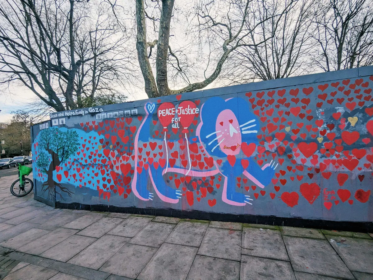 A long grey wall with a painting of a giant pink and blue lion. Red hearts of all sizes are painted all over. There is a painting of an olive tree on the left. The words read, "Peace + Justice for all."