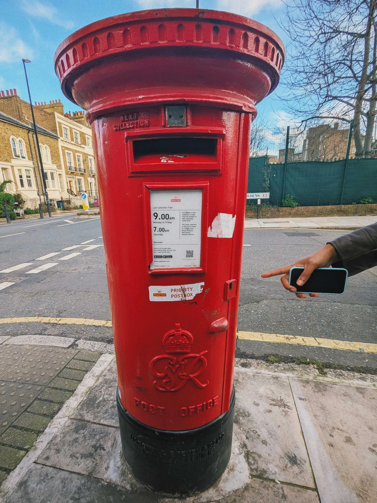 A bright red postbox with the initials GvR embossed on the front. K is pointing at it while holding her phone.