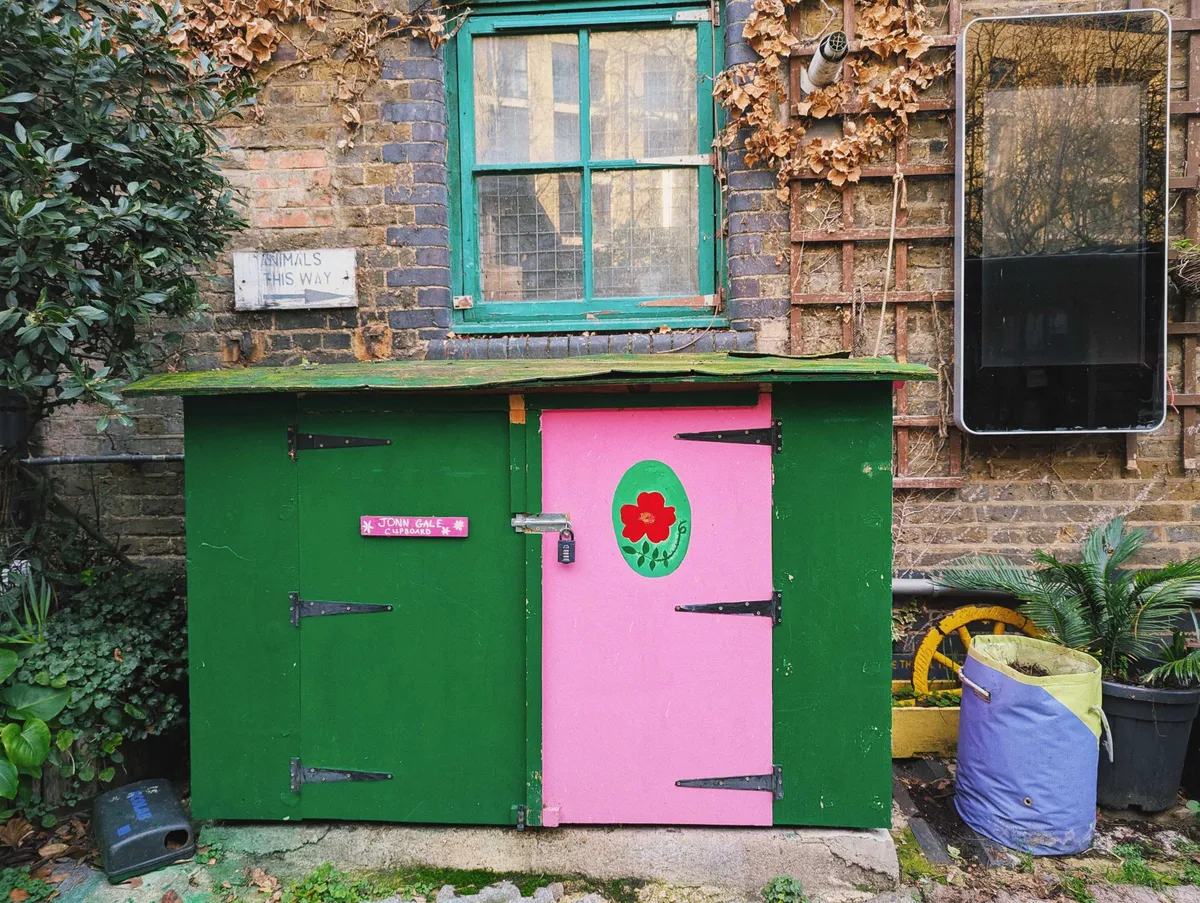 A small shed that's been painted green. The door is bright pink with a painting of a red flower inside a bright green oval.