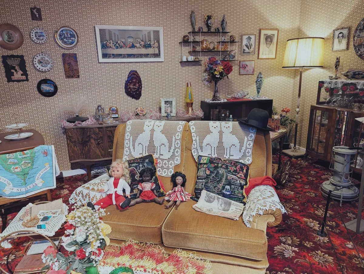 A 70s-looking living room with red floral carpeting and a velvet yellow couch. There are dolls and pillows stacked on the couch, with a pair of cat doilies laid across the top of the couch. There are a lot of things in this room.