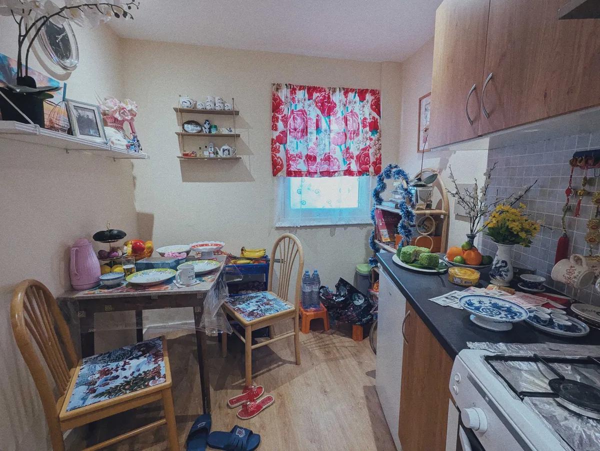 A dining nook within the kitchen. The table is covered with plates and has a clear plastic tablecloth. The stove has foil under the burner grates. There are house slippers on the floor.
