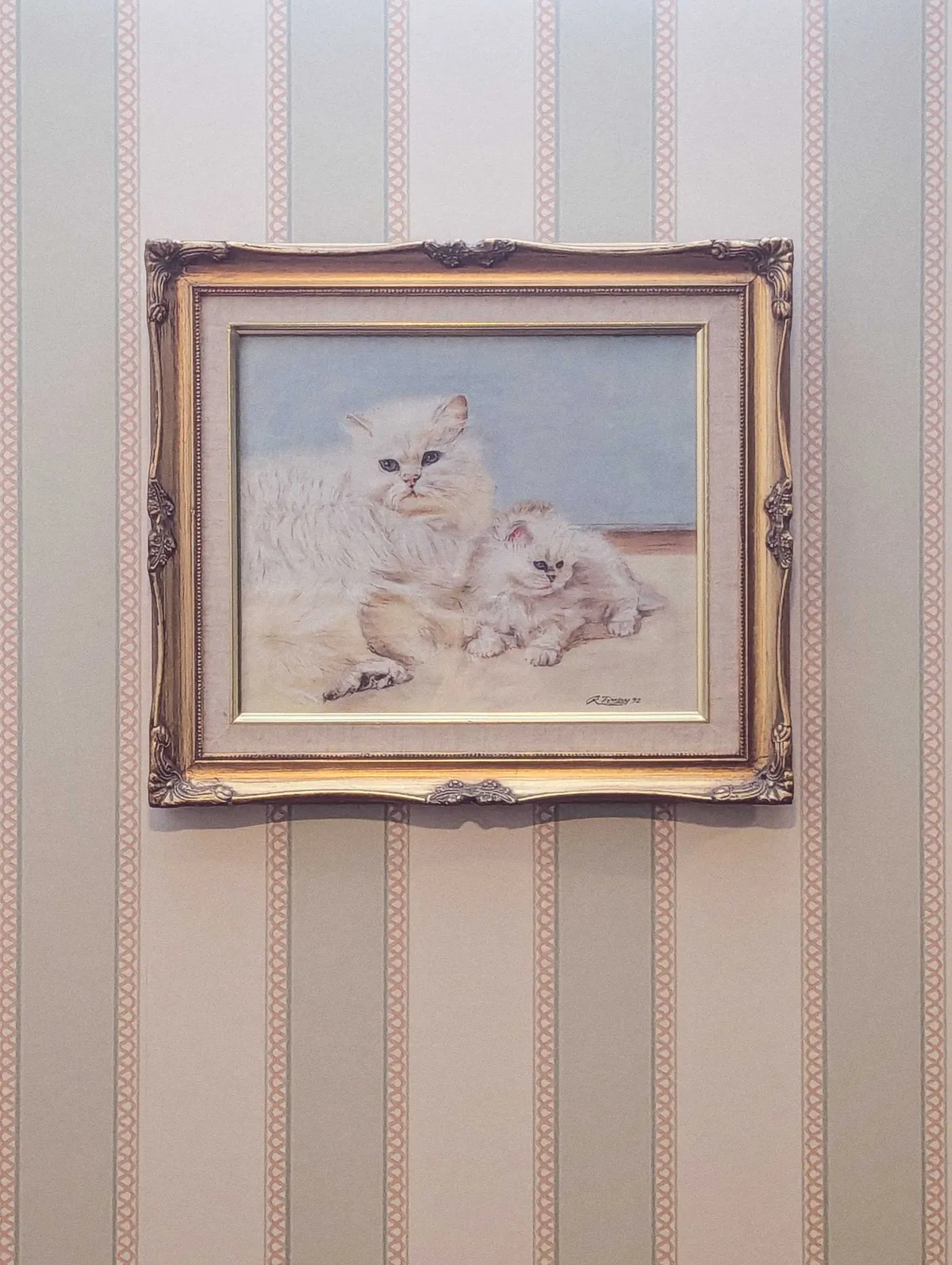 A painted portrait of two fluffy white cats set inside a golden frame. It's hung up against a striped, blue and white wallpaper background.
