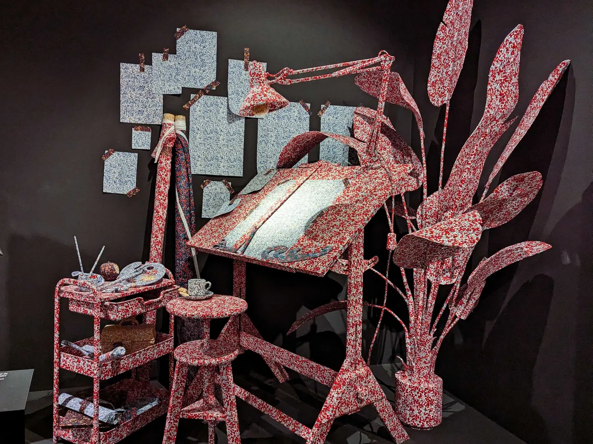 A textile designer's workstation that has been completely wrapped in a red patterned fabric, from the desk, to the stool, to the plant.