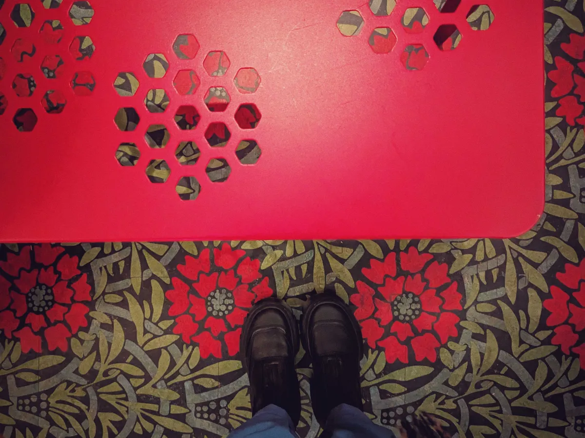A bright red bench with a perforated motif that sort of looks like a beehive pattern. The floor has a floral pattern with bright red flowers (perhaps poppies) against rich green leaves.