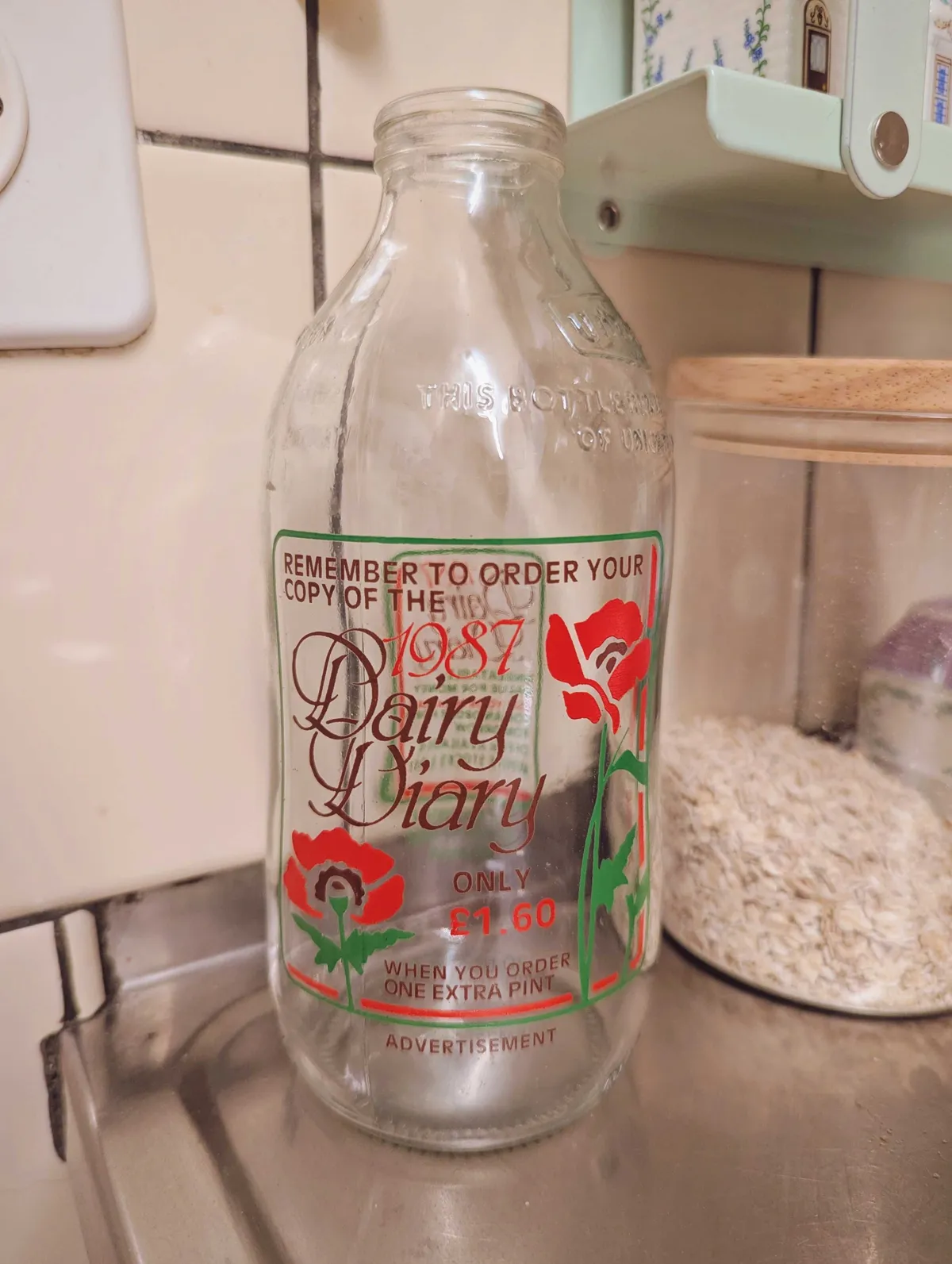 A clear, empty milk bottle with a green, red, and burgundy color palette. The text reads, "Remember to order your copy of the 1987 Dairy Diary/Only £1.60 when you order one extra pint/Advertisement." The text is housed within a green box with two poppies printed on the bottom.
