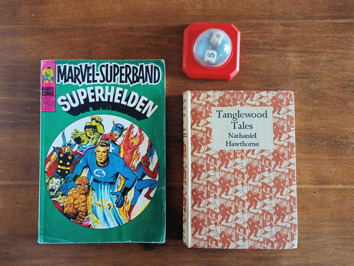 Comic book titled, "Marvel-Superband/Superhelden" with illustrations of marvel heroes on the cover; a red squarish object with a clear bubble top that contains a pair of die inside; a hardcover book titled, "Tanglewood Tales" with an allover pattern in white and rusty orange.