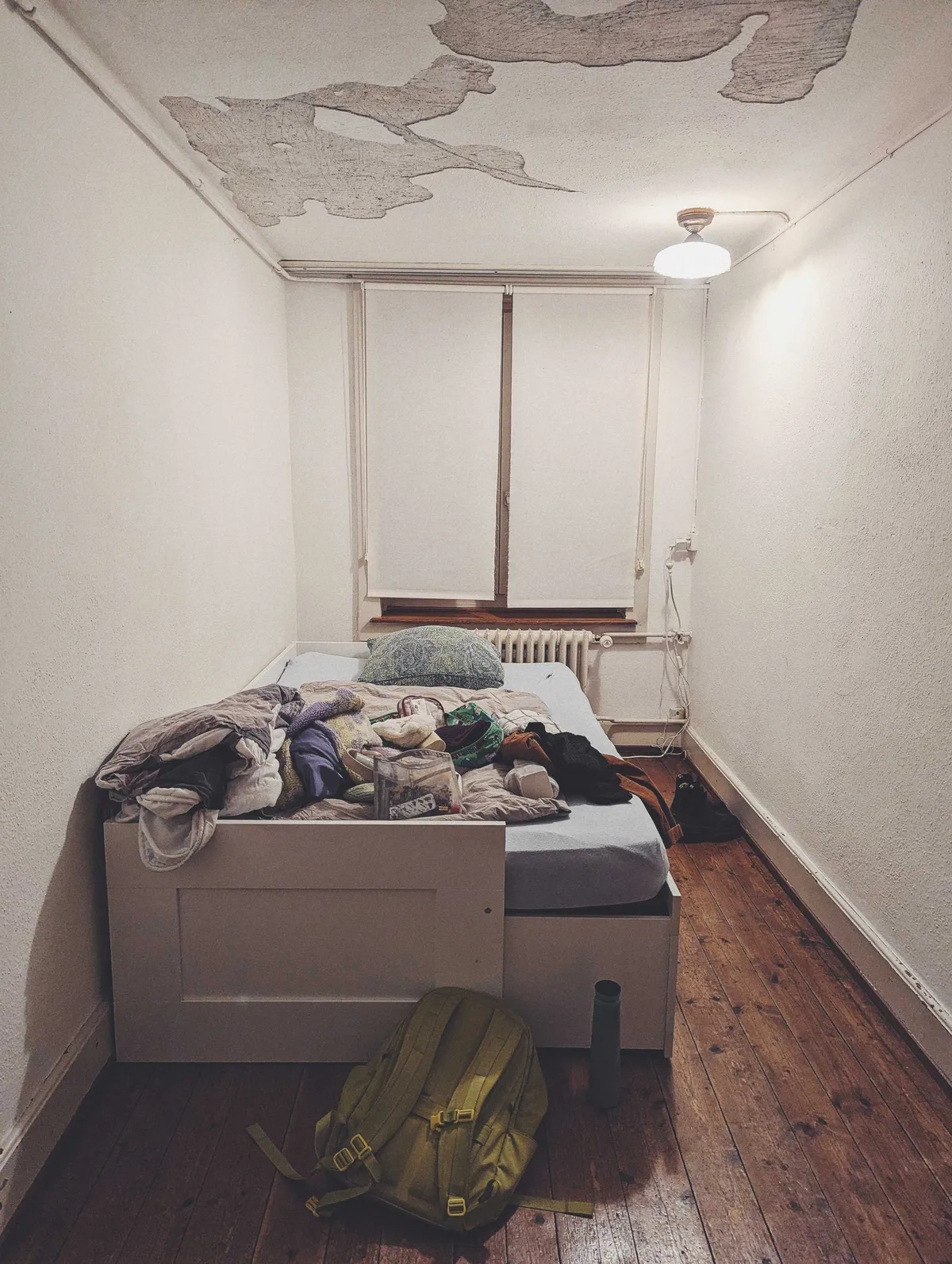 A small room with a bed in one corner. The bed is covered with items, mostly closed. There is a green backpack by the foot of the bed. The blinds are drawn shut.