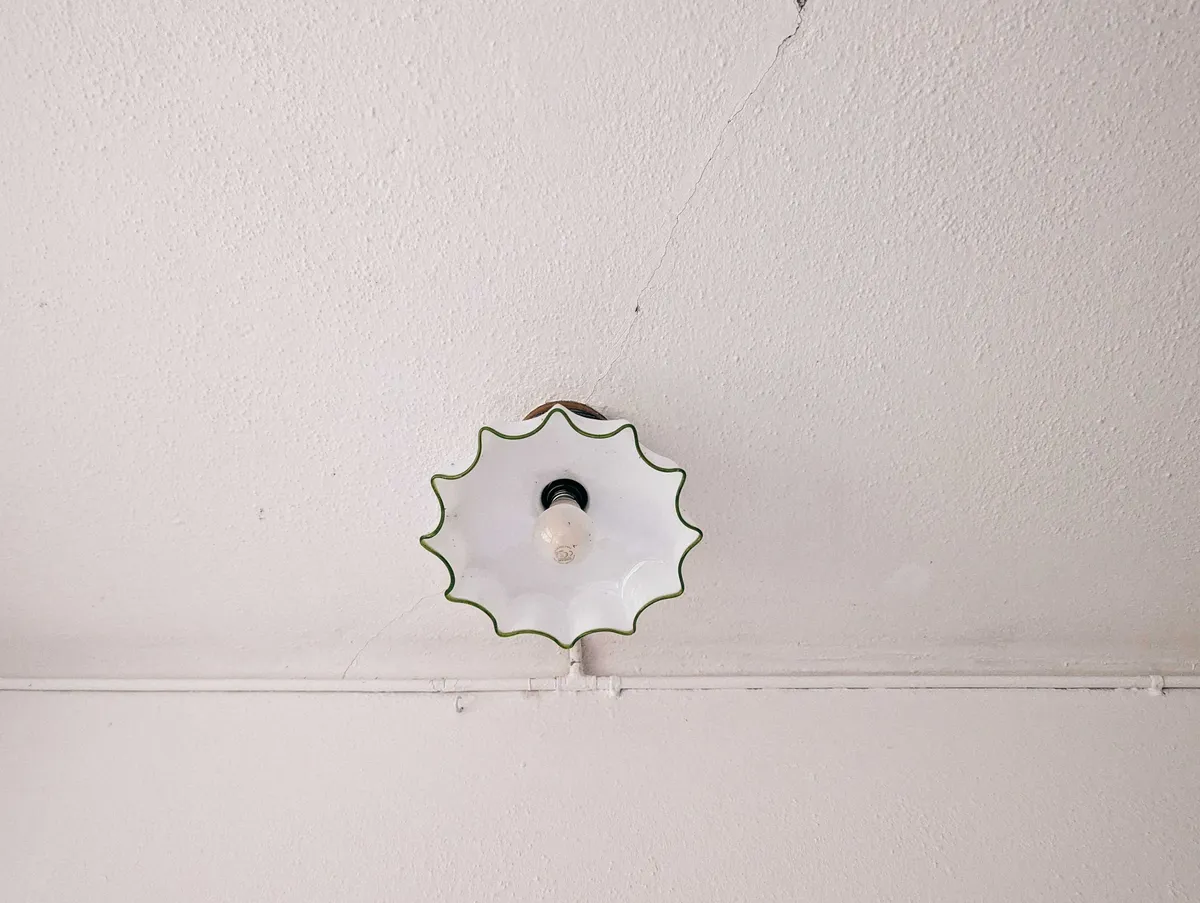 A white ceramic ceiling lamp with wavy edges lined in dark green against an off-white and old ceiling.