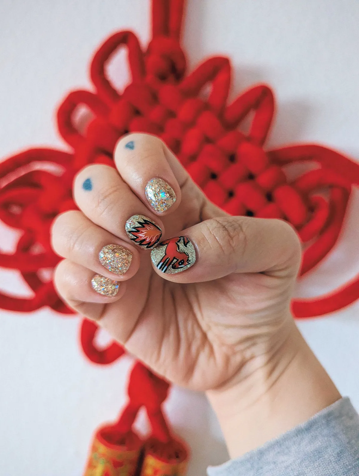 Peige's nails painted with gold glitter. The middle finger has flames painted on. The thumb is painted with a red horse with white feet.
