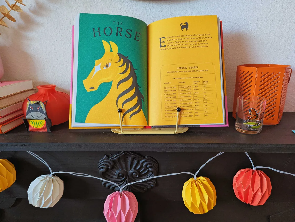 A black origami horse is leaning against a red vase shaped like a squat lantern. To the right right is an open spread with a yellow horse against a green background. To the right of that is a shotglass with a brown horse painted on it. Behind that is an orange metal lantern.