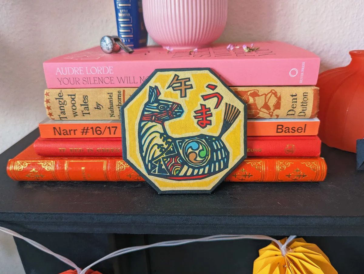 A painting of a horse on a hexagon shaped coaster. It is leaning against a stack of books that are pink, red, and orange toned.