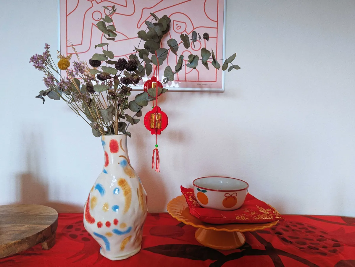 Dried flowers in a colorful, handmade vase. There are red lanterns hanging from the eucalyptus. An orange cake stand with a bowl decorated with oranges on a red puffy pillow is to the right of the vase.