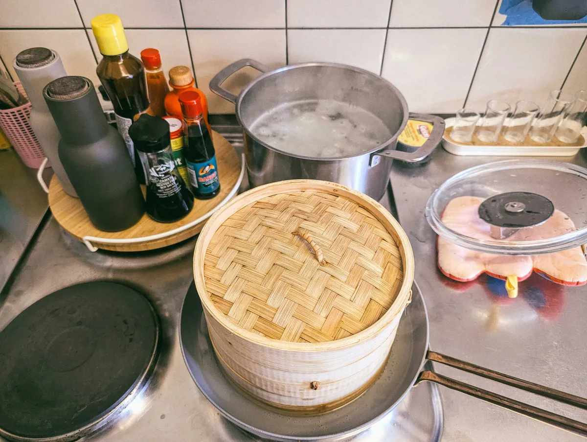 A bamboo steamer cooks on top of a grey pan on the stove. There are various sauces on the left and a pot of boiling water on the back burner.