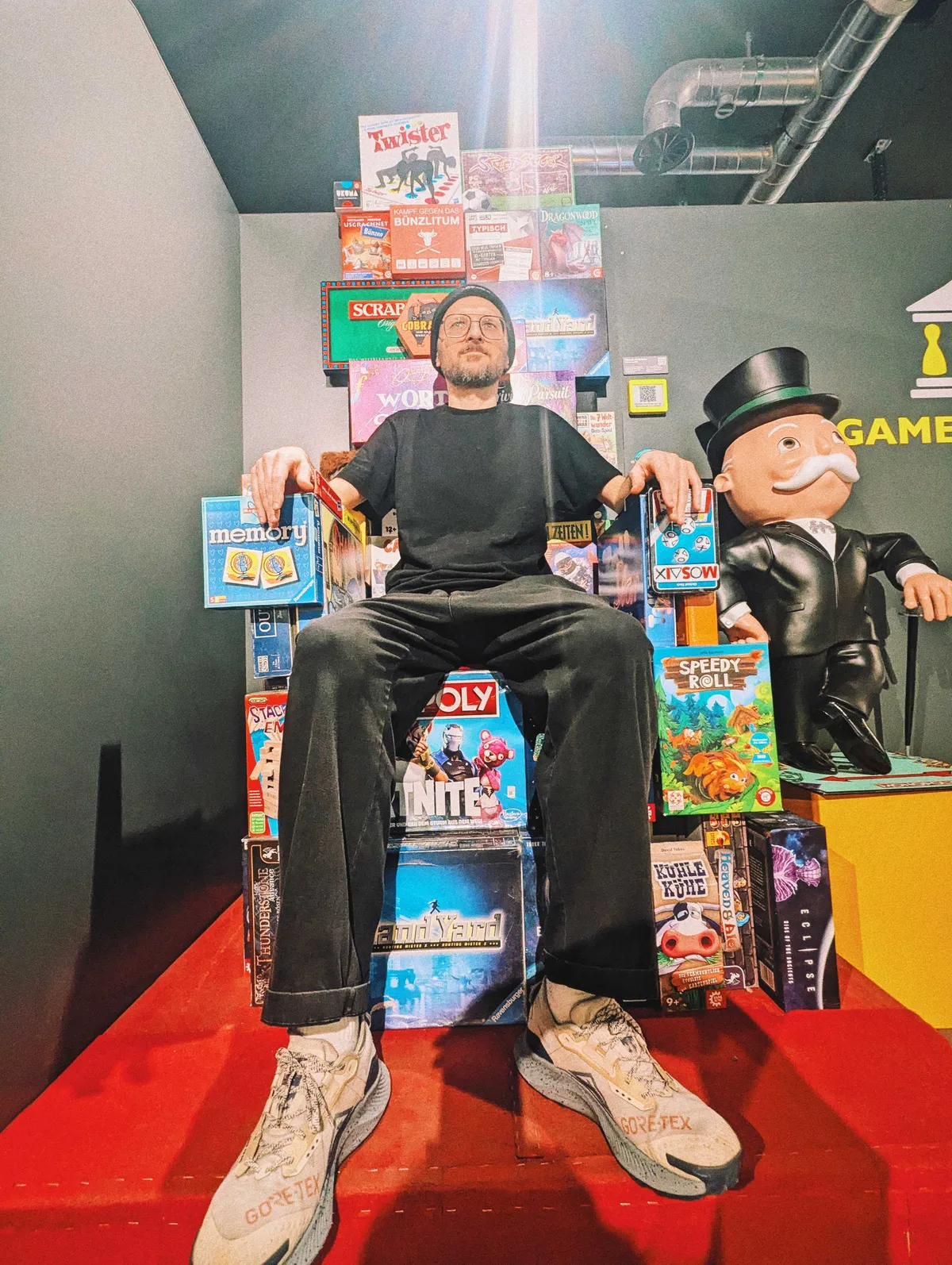 A white man with glasses and a beard wearing all black sits on a throne made up of board games.