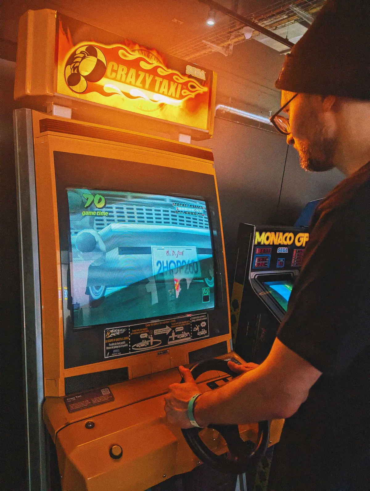 Daniel standing with his hands on the wheel of the arcade machine. It's yellow with a glowing "Crazy Taxi" sign inside a flaming fireball.