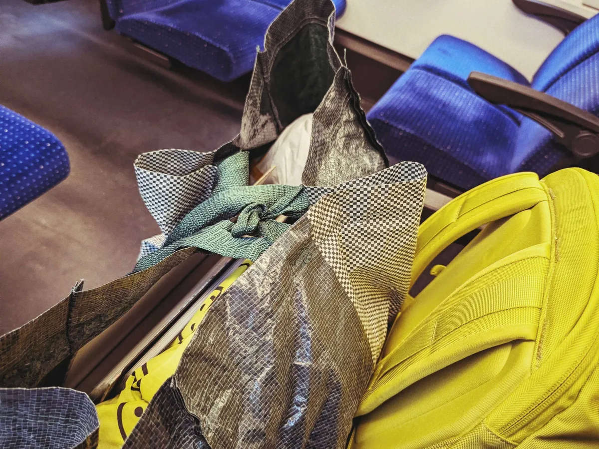A dark green patterned Ikea bag (from the Hay collab) filled with items, next to a lime green backpack, both on a train seat.