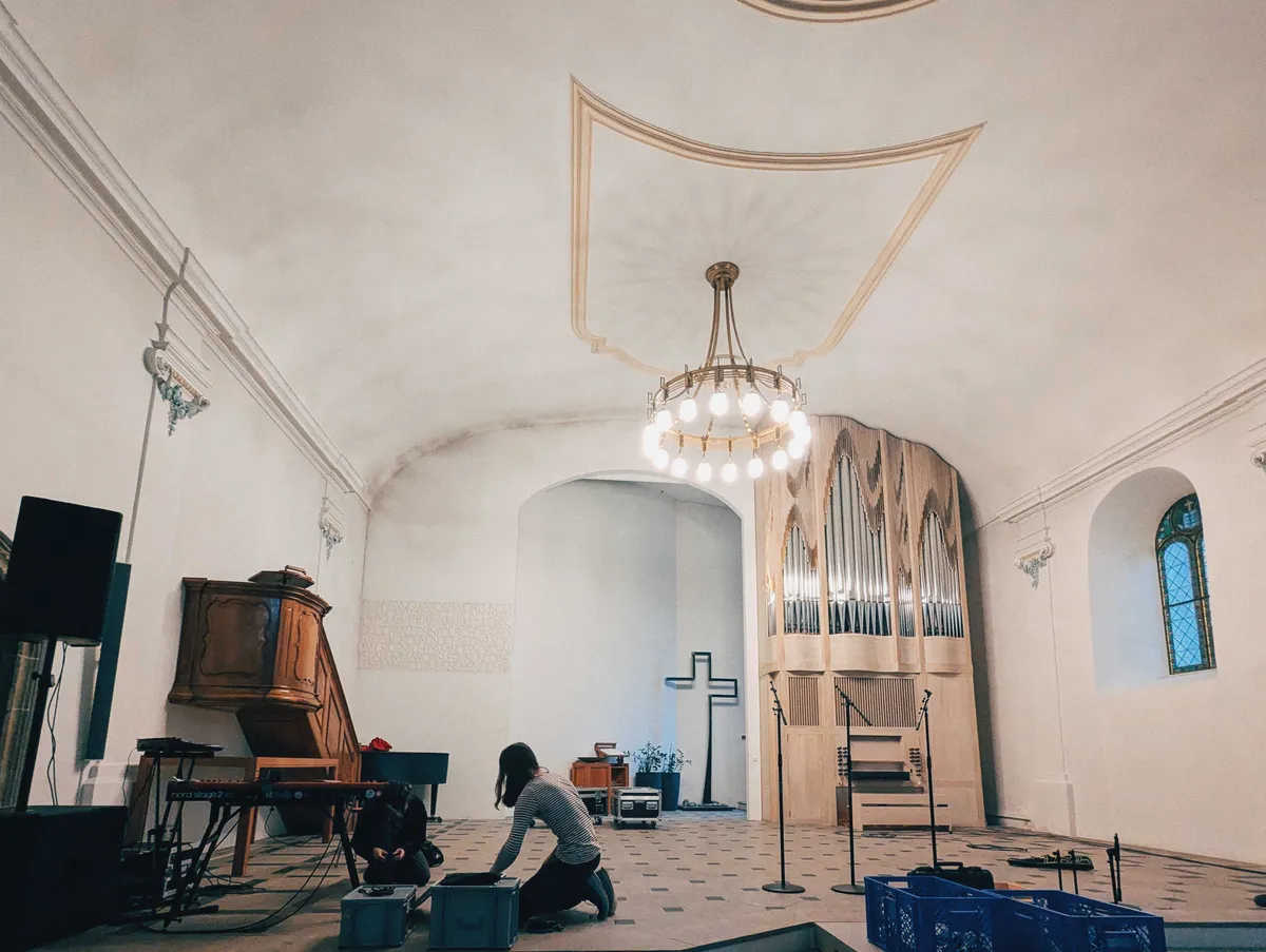 Two people bent over and putting together cables nears the floor. The inside of the church is white with high ceilings. A modern organ is built into the right back corner of the wall. Next to that is a large, modern-looking, metal cross.