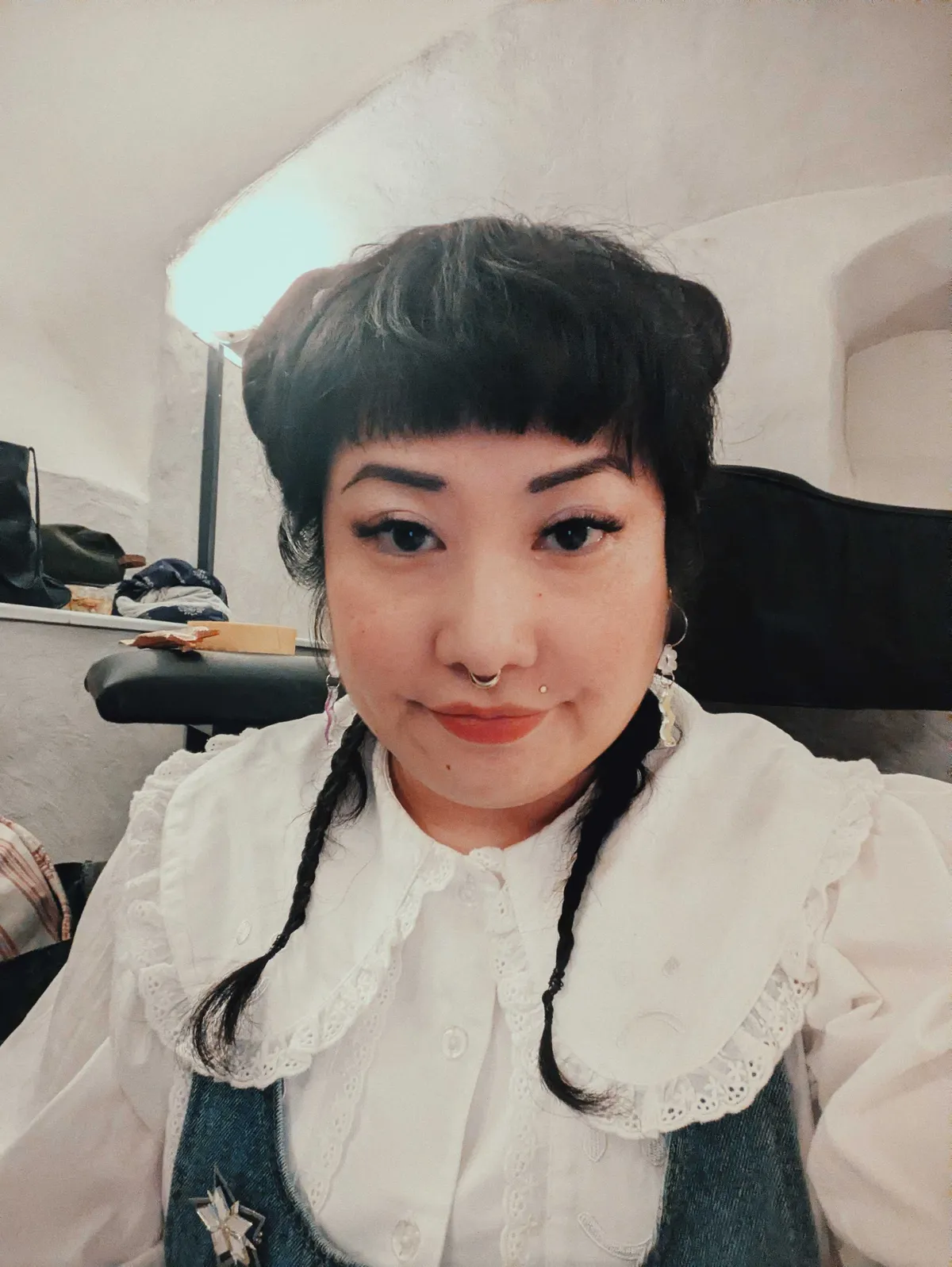 Peige staring into the camera with a slight smile. They have buns peeking out from the top of their head and two small braids falling down the front of their chest.