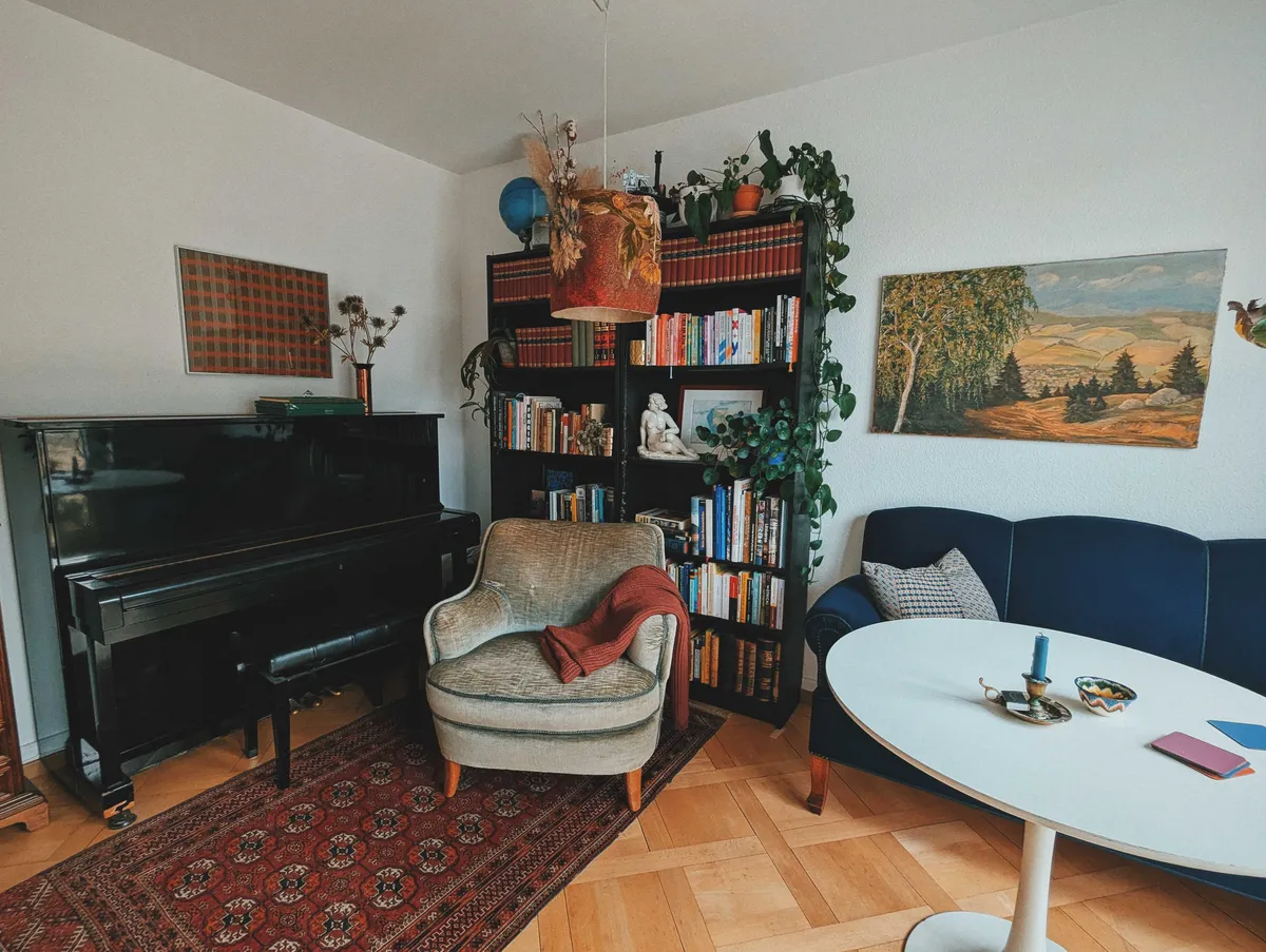 A corner of the living room with a bookshelf filled with books, a black upright piano, a Turkish rug, and plush seating areas. There is a framed checkered pattern hanging above the piano, and landscape painting above the dark blue sofa.