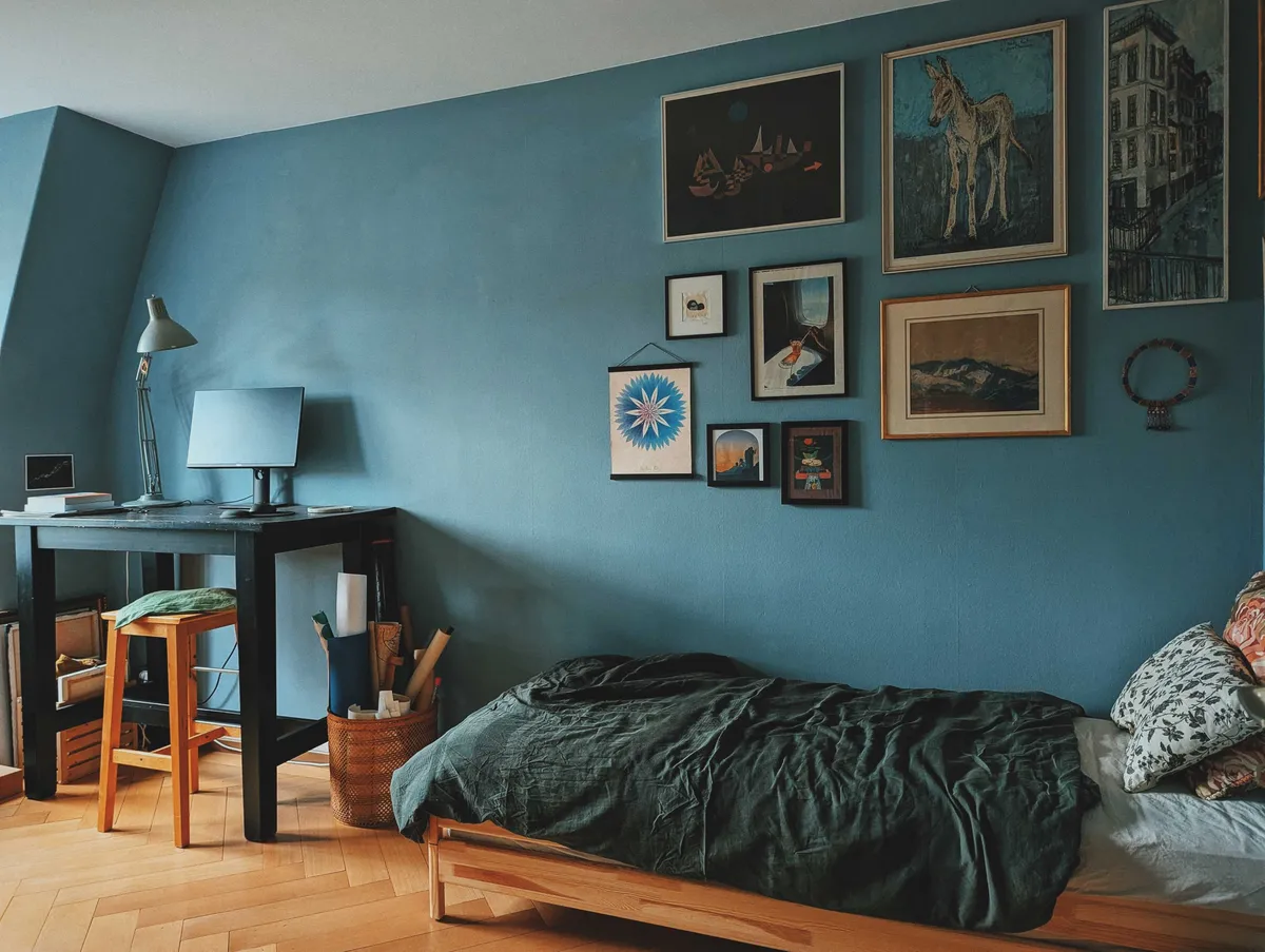 A room with dark turquoise walls. A gallery of paintings hangs above the bed but have been arranged so they only fill up the right corner of the wall. There is a standing desk with a computer at the foot of the bed.