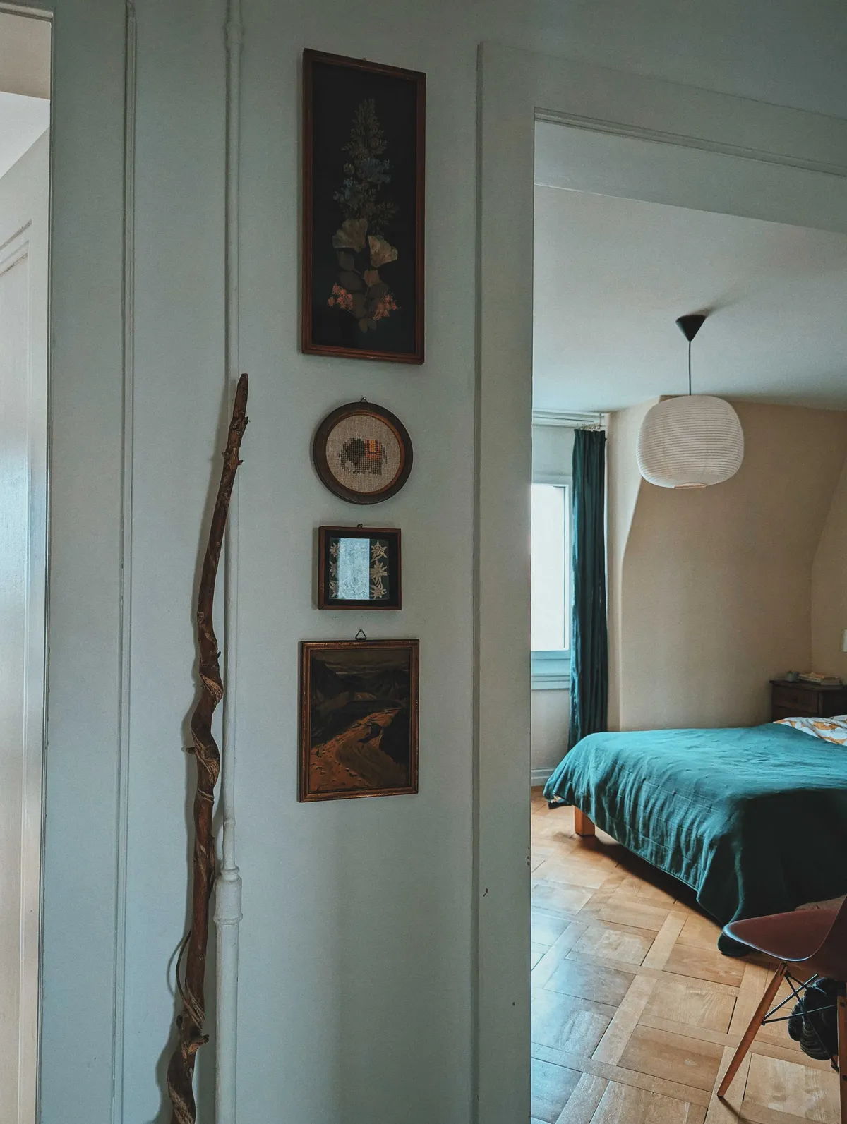 A corner of the hallway showing a vertical row of small art hanging from the wall. Next to it is a long stick with some twisting roots leaning against the wall. You can see a glimpse of K's bedroom, with parquet flooring and dark turquoise bedsheets peek into view.
