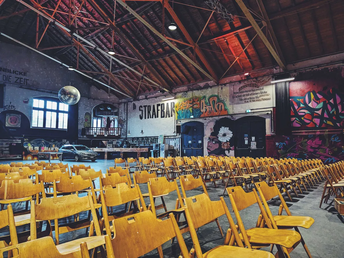 Rows of wooden folding chairs in the foreground. The room has high wooden ceilings and the walls are painted with different styles of street art.