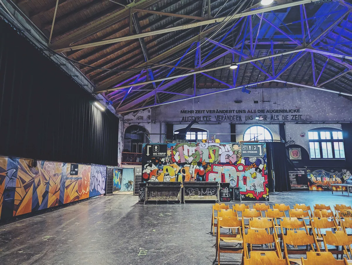 Another angle of the room, showing some chairs in the foreground, the scuffed, black cement floors, and a container in the back that has been painted with layers of graffiti.