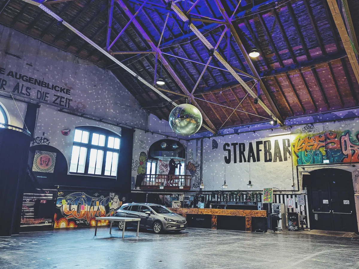 A hatchback car is parked next to the bar. It looks like the size of a couch in this large space. There is a giant disco hanging from the ceiling and speckles the walls with light, even during the day time.