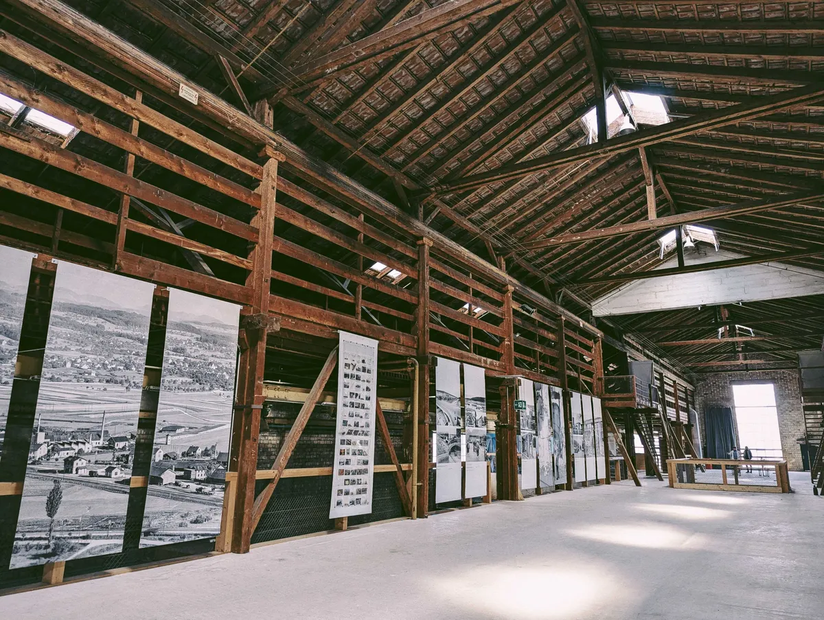 A long room with high wooden ceilings and white cement walls. There are wooden beams acting as walls, where large prints containing information and pictures of the space have been hung.