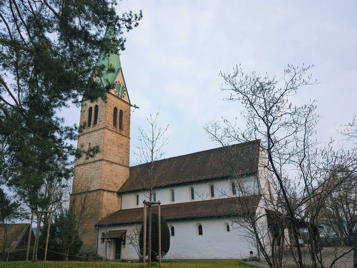 A white, old building with tapered brown roofs and tiny arched windows. There is a tall bell tower made of red brick. The spire is bright green from oxidization, matching the green of the clock face.