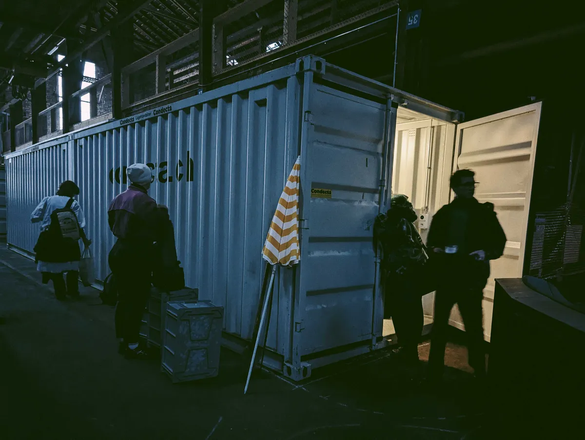 A large, white shipping container with one door open, emitting light from the inside. There are two people standing by the doorway, and two more towards the side of the container.