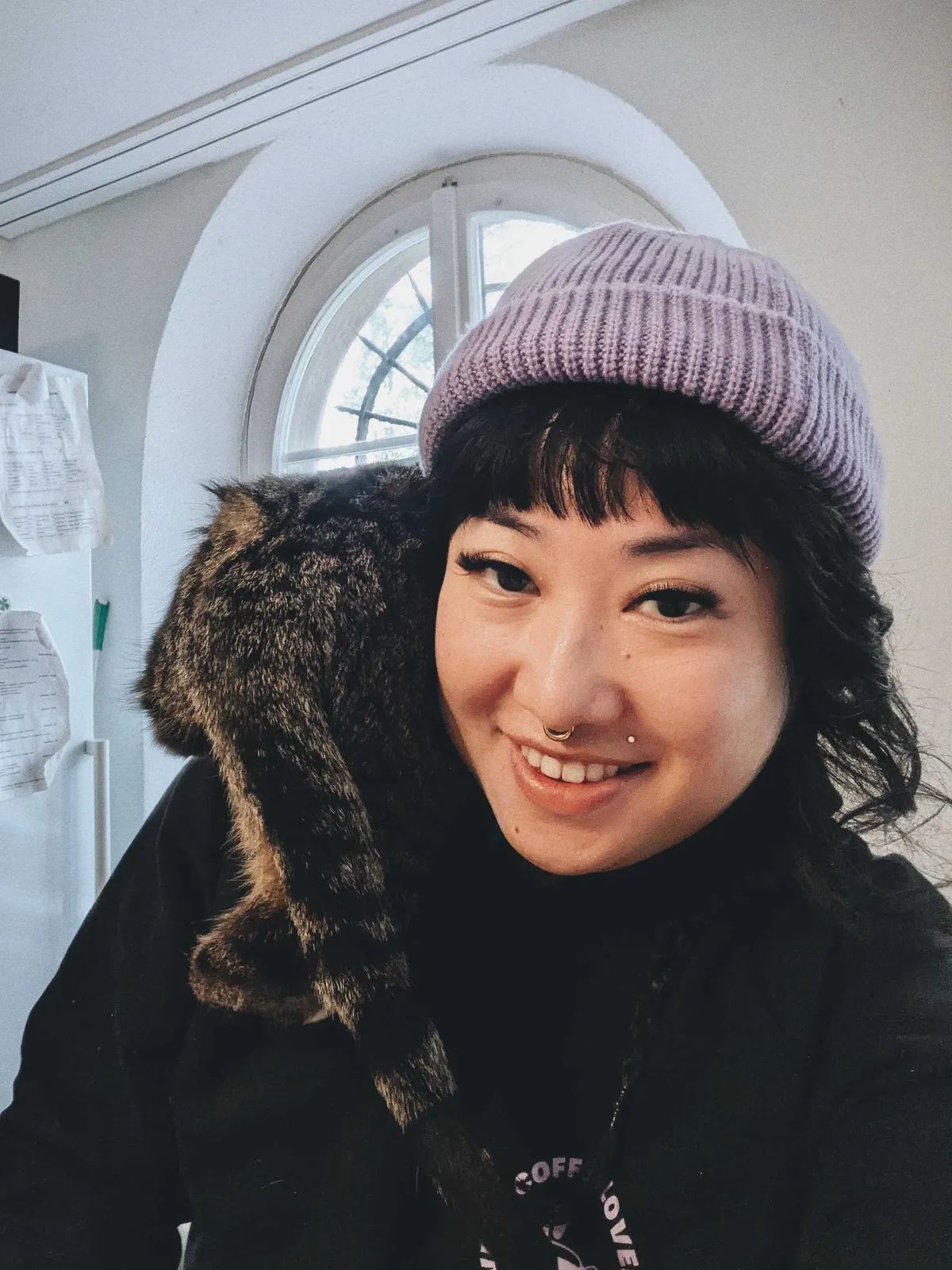 Peige, leaning slightly forward and smiling at the camera. Neptune the cat is perched on their shoulder, his back facing the camera.