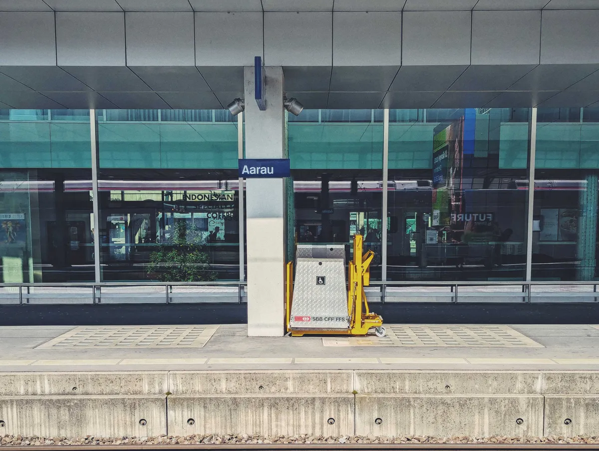 The train platform across the tracks, with giant tinted windows of the station in the background. There's a blue sign with white type that says, "Aarau."