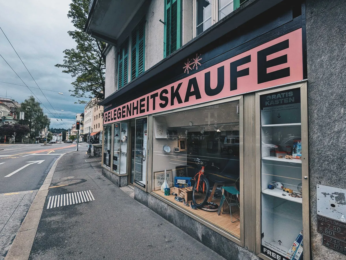 View of the shop window at a slight angle. There are lots of antique-looking items in the window. The name is in bold, black and uppercase text against a pink background.