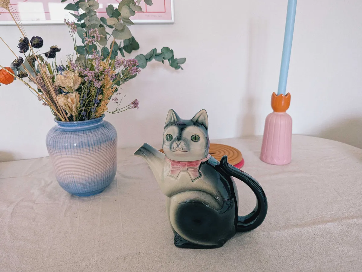 A teapot in the shape of a cat wearing a pink ribbon around its neck.