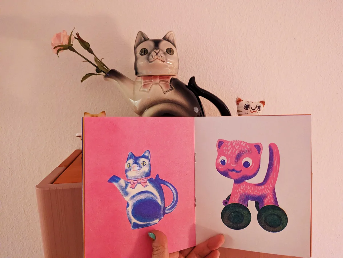 Peige holding up a zine that has the exact image of the carafe cat wearing a pink ribbon with the derpy face.