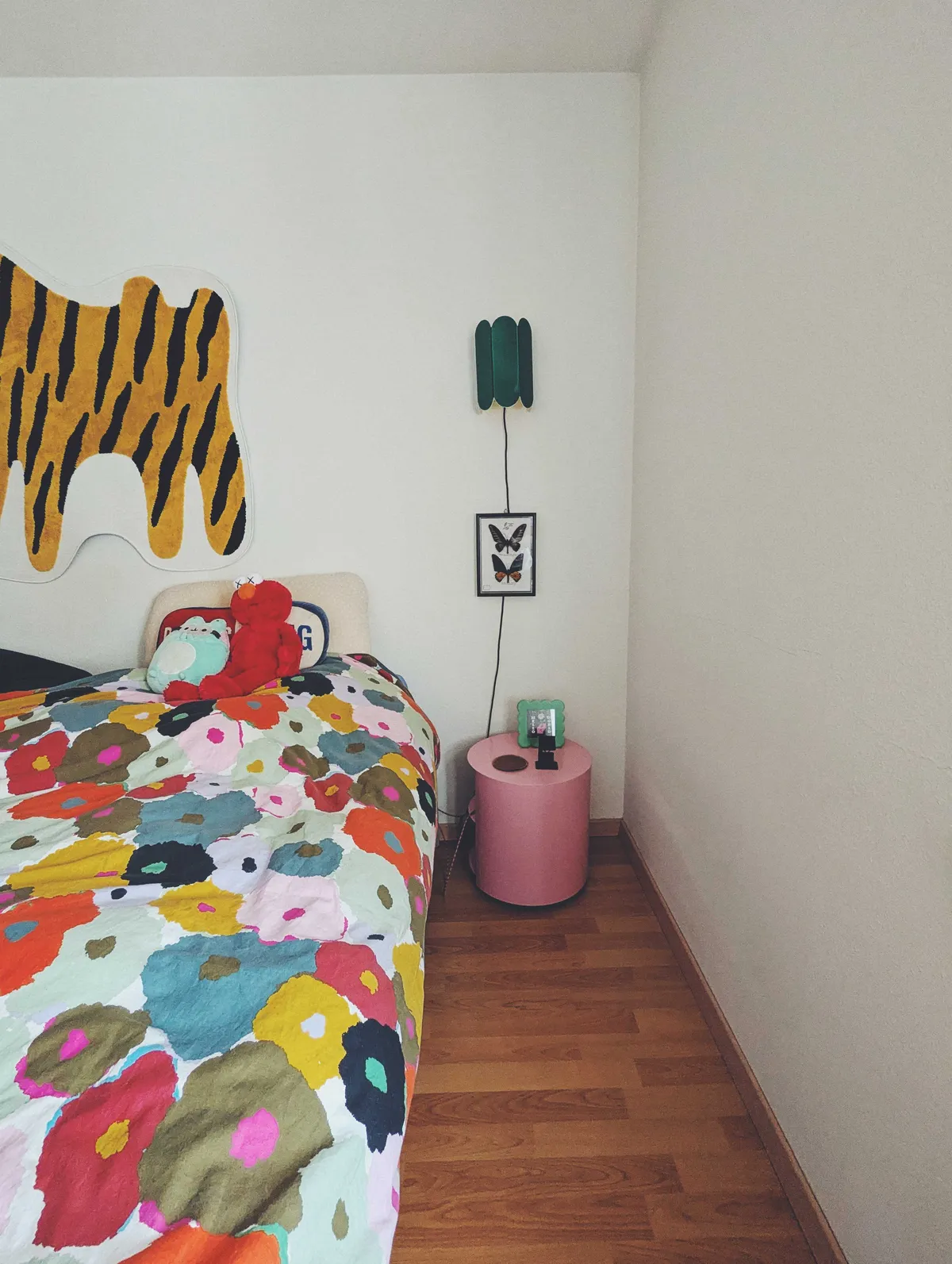 A clean bedside area with the rug removed, revealing wooden floors. The bed is made and decorated with some plushies.