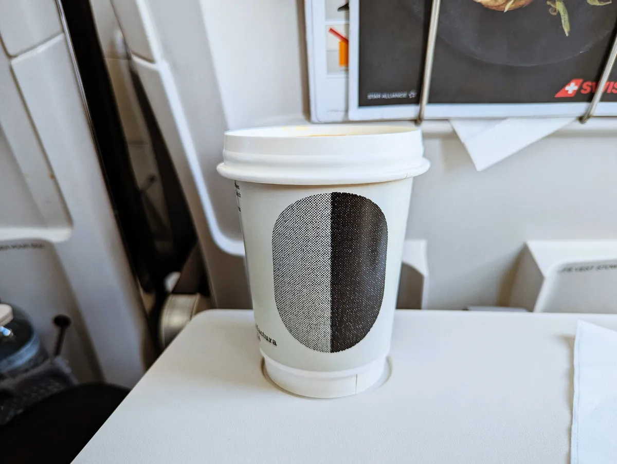 A cup of coffee with Miró's signature bean logo on the cup. The cup sits on the airplane fold-out tray.