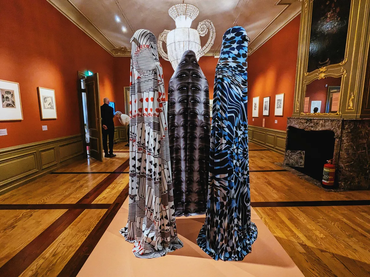 Three towering mannequins stand back-to-back with each other, each fully covered in fabric of varying patterns.