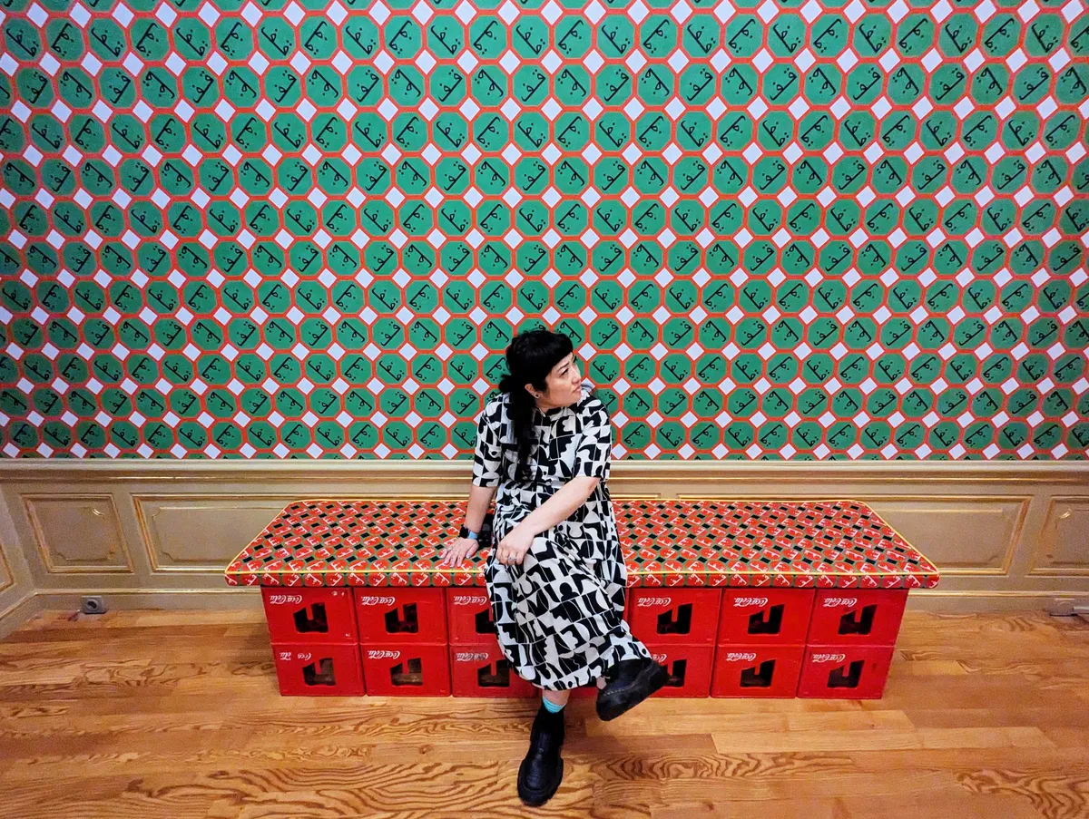 Peige sitting on red Coca Cola crates on a cushion of red, green, and white Arabic-inspired patterns. The wallpaper is mostly green with some red and white, and black Arabic writing.
