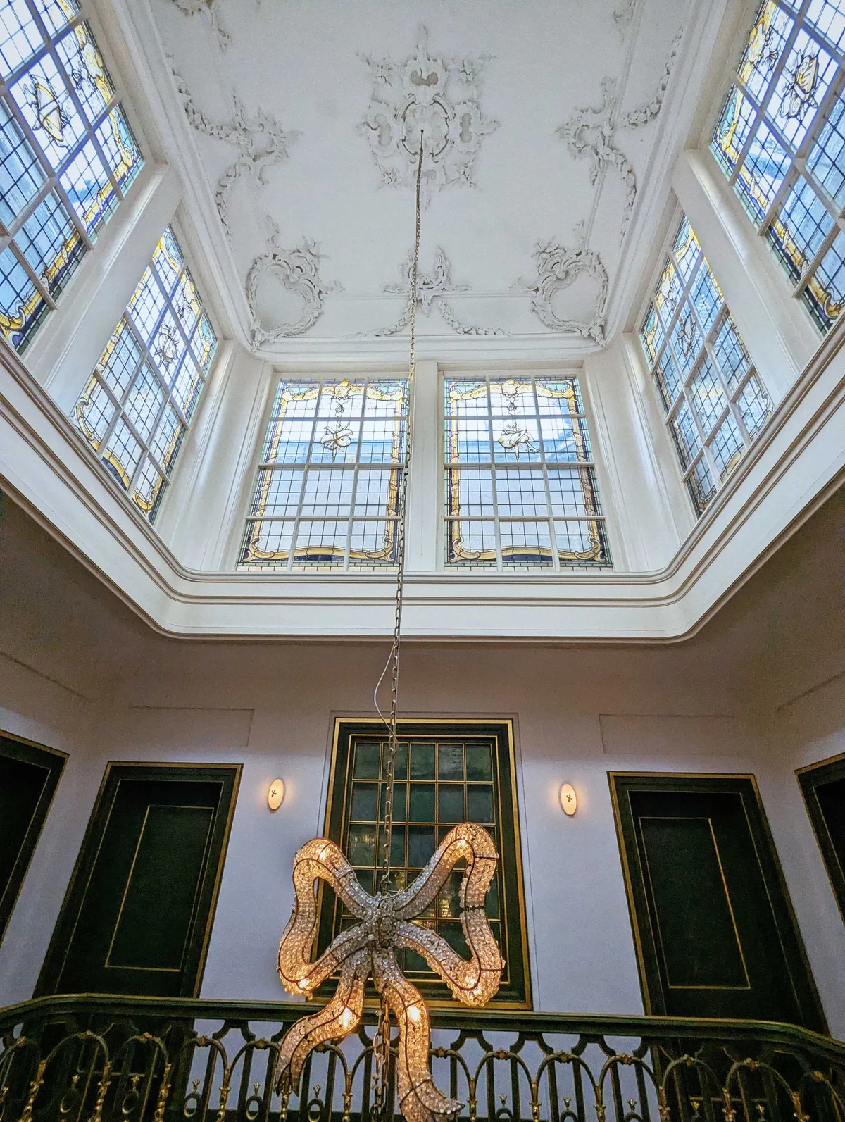 Chandelier in the shape of a bow, hanging from an ornate ceiling with stained glass windows.